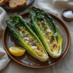 Two halves of grilled romaine lettuce topped with herbs, onions, and lemon on a brown plate.