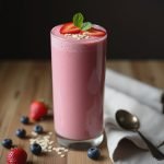 Tall glass of pink smoothie topped with strawberries and mint leaves, surrounded by berries and oats.